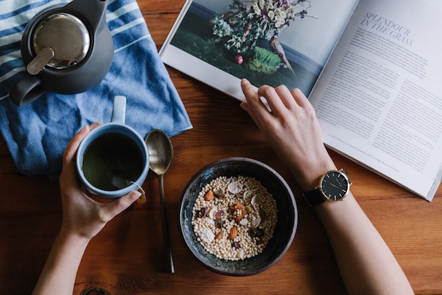 A bowl with food in front of someone holding a cup of coffee and flipping through a book.