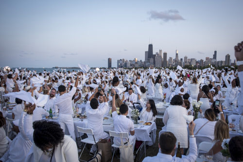 Hundreds of diners, seated at banquet tables, each wearing all white, are cheering and celebrating at an outdoor event with the Chicago Skyline in the background.
