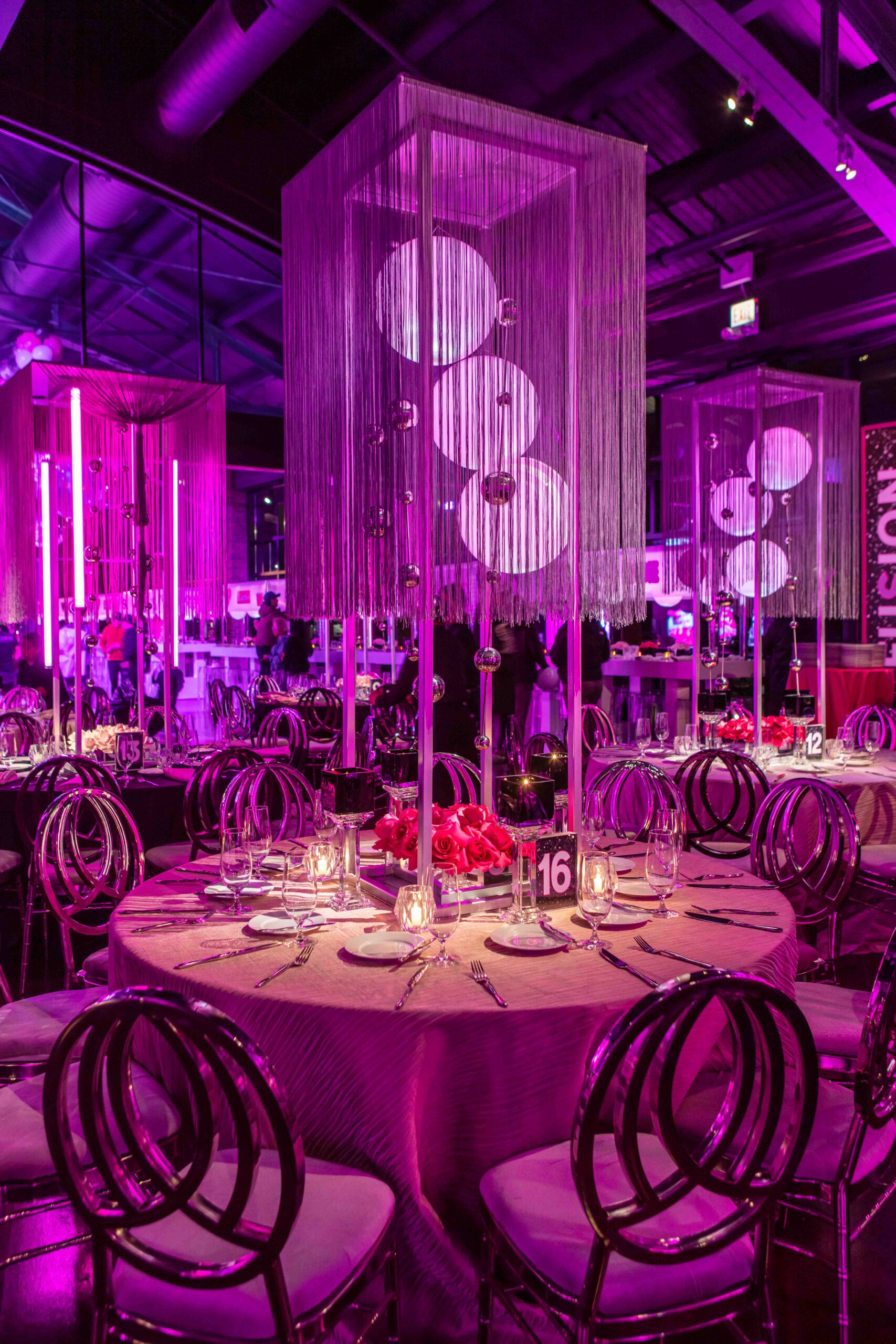 Magenta-themed event space, focusing on a table bathed in golden light, surrounded by glossy gold chairs, with magenta neon lights and unique chandeliers.