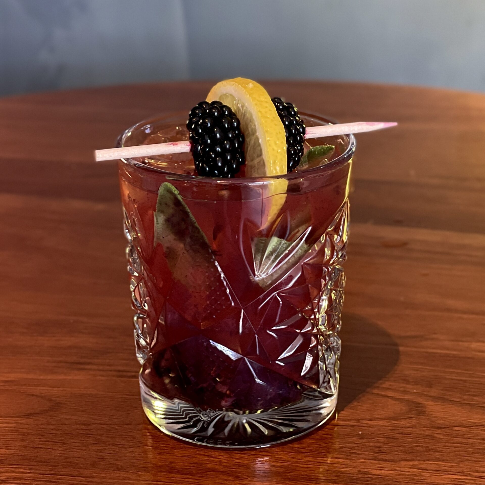 A cocktail in a crystal rocks glass, garnished with a blackberry and lemon skewer.