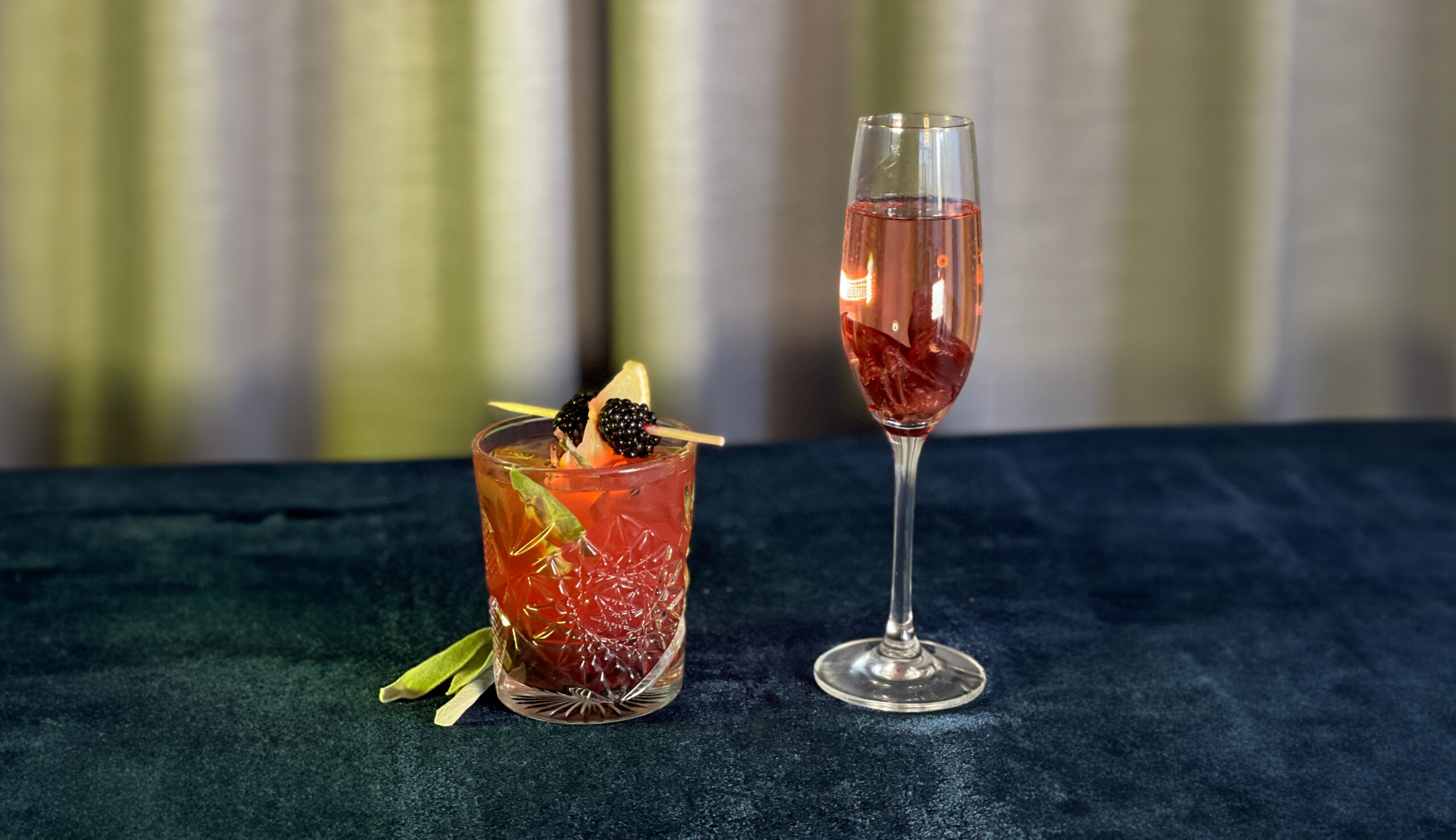 Two cocktails, side by side. One in a rocks glass garnished with a blackberry and lemon skewer, and one in a champagne flute, garnished with an edible hibiscus flower.